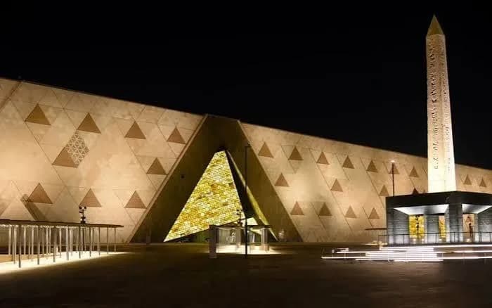 Exterior of the Grand Egyptian Museum illuminated at night, with an ancient obelisk in front