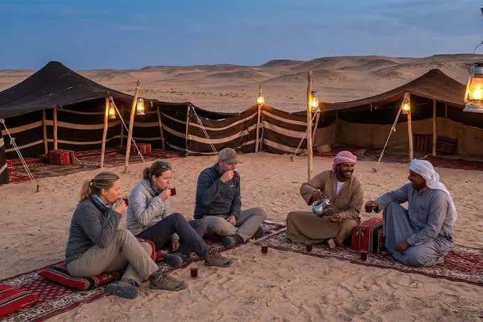 Guests seated on cushions at a desert camp in the evening, with lanterns lit